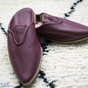May include: A pair of burgundy leather slippers with pointed toes. The slippers have a simple design and are made of soft, supple leather.