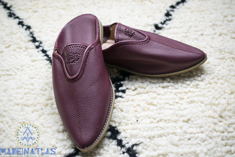 May include: A pair of burgundy leather slippers with pointed toes. The slippers have a simple design and are made of soft, supple leather.