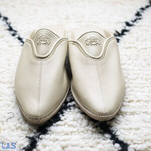 May include: A pair of gold leather slippers with a pointed toe and a closed back. The slippers are on a white fluffy rug.