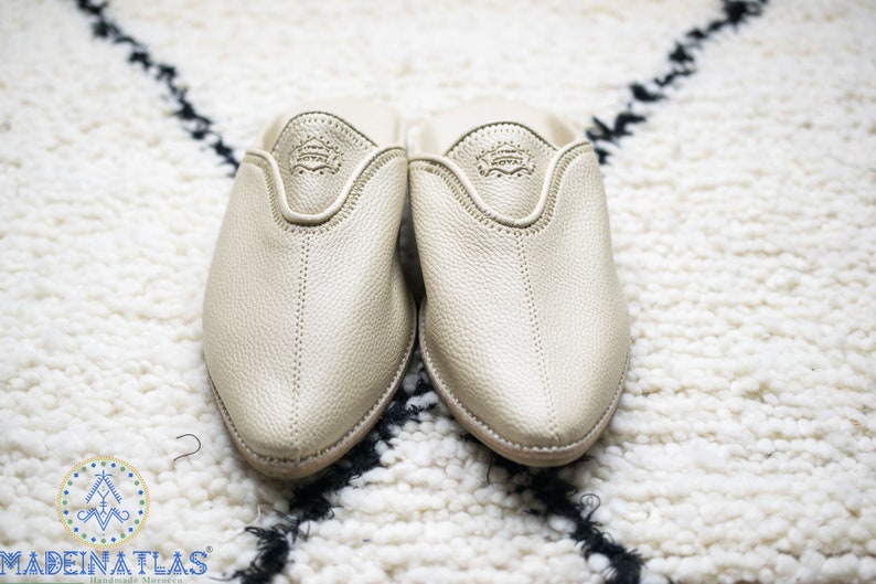 May include: A pair of gold leather slippers with a pointed toe and a closed back. The slippers are on a white fluffy rug.