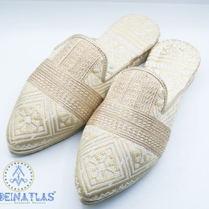 May include: Pair of gold embroidered slippers with a pointed toe and a woven strap across the top. The slippers have a white background with a gold geometric pattern.