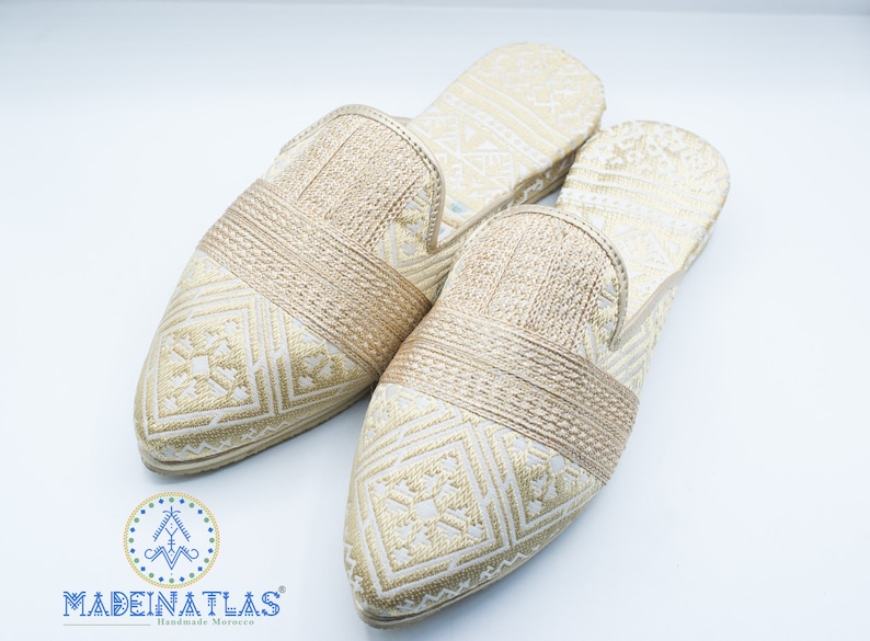 May include: Pair of gold embroidered slippers with a pointed toe and a woven strap across the top. The slippers have a white background with a gold geometric pattern.
