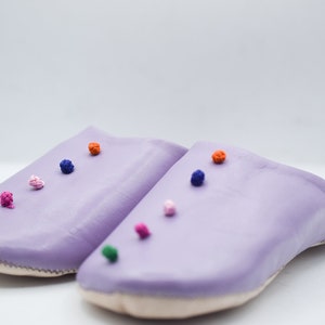 Op de afbeelding: Een paar lavendelkleurige leren slippers met kleurrijke pompons. De slippers hebben een ronde neus en een gestikte naad rond de rand. De pompons zijn in tinten groen, roze, oranje, blauw en wit.