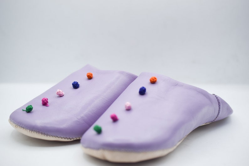 Op de afbeelding: Een paar lavendelkleurige leren slippers met kleurrijke pompons. De slippers hebben een ronde neus en een gestikte naad rond de rand. De pompons zijn in tinten groen, roze, oranje, blauw en wit.