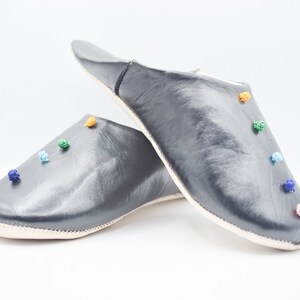 Op de afbeelding: Een paar donkergrijze leren slippers met kleurrijke pompons. De slippers hebben een ronde neus en een zachte, cr&egrave;mekleurige zool. De pompons zijn in tinten roze, blauw, groen en oranje.