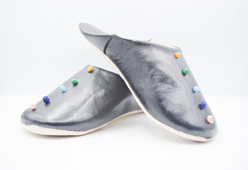 Op de afbeelding: Een paar donkergrijze leren slippers met kleurrijke pompons. De slippers hebben een ronde neus en een zachte, cr&egrave;mekleurige zool. De pompons zijn in tinten roze, blauw, groen en oranje.
