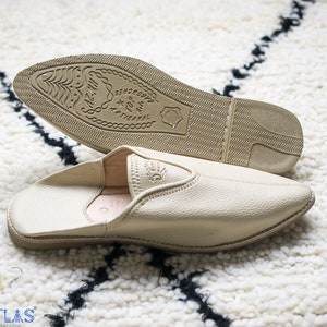 May include: A pair of beige leather slippers with a pointed toe and a closed back. The slippers have a rubber sole with the text "100% ARTISANAL" printed on the bottom.