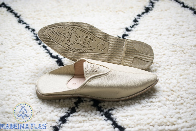 May include: A pair of beige leather slippers with a pointed toe and a closed back. The slippers have a rubber sole with the text "100% ARTISANAL" printed on the bottom.