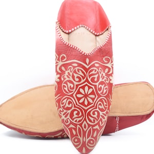 Puede incluir: Un par de zapatillas de cuero rojas con un patrón floral blanco. Las zapatillas tienen una punta puntiaguda y una suela plana.