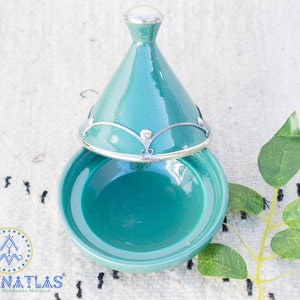 May include: A teal ceramic tagine with a silver lid and decorative metal accents. The tagine is open and the inside is visible.