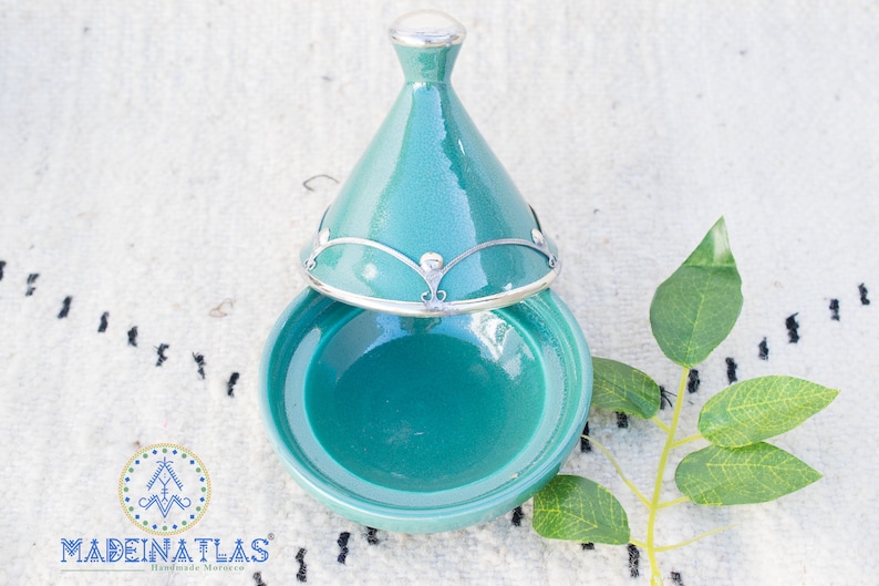 May include: A teal ceramic tagine with a silver lid and decorative metal accents. The tagine is open and the inside is visible.
