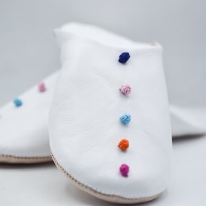 Op de afbeelding: Een paar witte leren slippers met kleurrijke pomponaccenten. De slippers hebben een eenvoudig ontwerp met een ronde neus en een neutrale zool. De pompons zijn in tinten roze, oranje, blauw en paars.