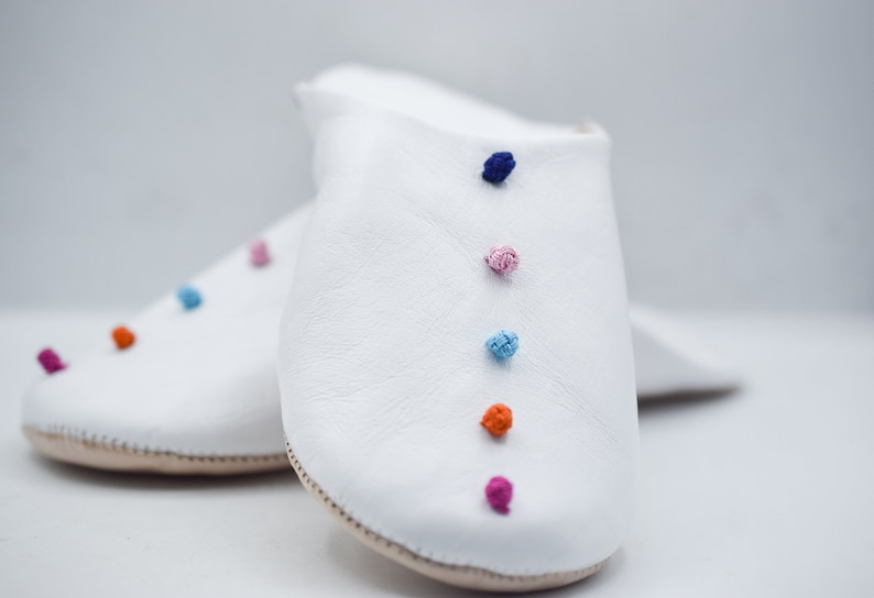 Op de afbeelding: Een paar witte leren slippers met kleurrijke pomponaccenten. De slippers hebben een eenvoudig ontwerp met een ronde neus en een neutrale zool. De pompons zijn in tinten roze, oranje, blauw en paars.