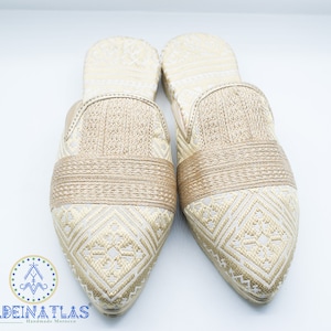 May include: Pair of gold and white embroidered slippers with pointed toes. The slippers have a woven band across the top.