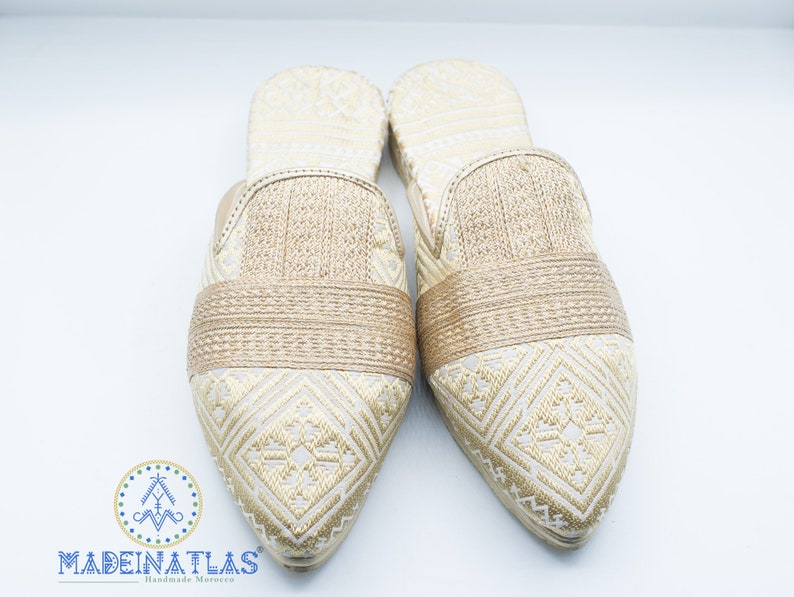 May include: Pair of gold and white embroidered slippers with pointed toes. The slippers have a woven band across the top.