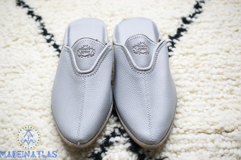 May include: A pair of gray leather slippers with pointed toes. The slippers have a simple design with a small embroidered logo on the top.