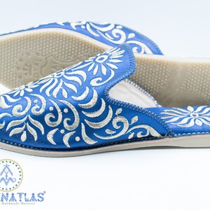 May include: A pair of blue embroidered slippers with a white floral pattern. The slippers have a pointed toe and a flat sole. The brand name "MADEINATLAS" is visible on the bottom of the slippers.