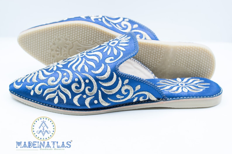 May include: A pair of blue embroidered slippers with a white floral pattern. The slippers have a pointed toe and a flat sole. The brand name "MADEINATLAS" is visible on the bottom of the slippers.