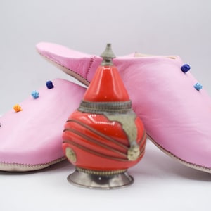 Op de afbeelding: Een paar roze leren slippers met kleurrijke pompons en een rode en zilveren decoratieve fles. De slippers zijn zachtroze en hebben een eenvoudig ontwerp. De fles is rood met zilveren accenten.