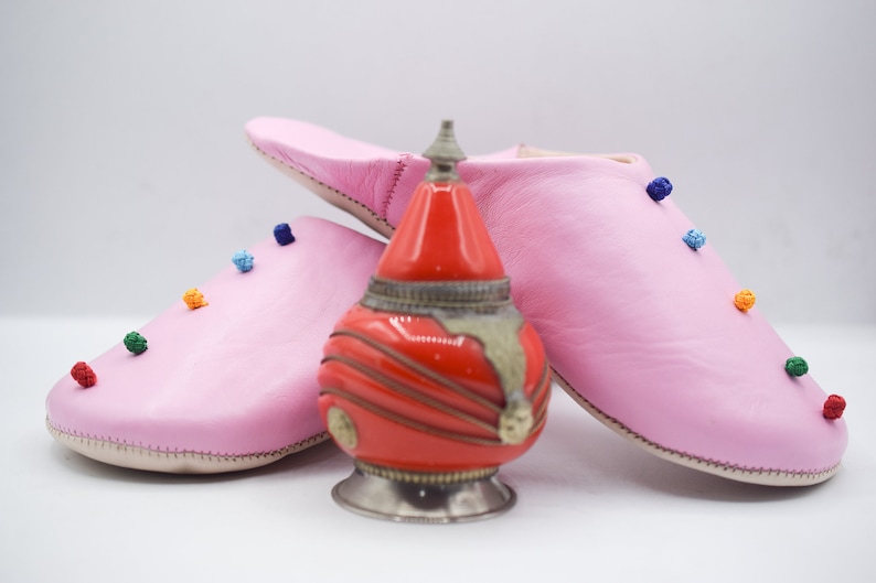 Op de afbeelding: Een paar roze leren slippers met kleurrijke pompons en een rode en zilveren decoratieve fles. De slippers zijn zachtroze en hebben een eenvoudig ontwerp. De fles is rood met zilveren accenten.