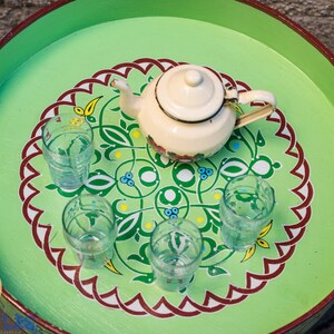 May include: A green tray with a white teapot and five clear glasses. The tray has a decorative design with red, white, and green colors.