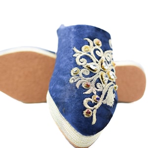 Puede incluir: Un par de zapatillas azules bordadas con hilo dorado y joyas de colores. Las zapatillas tienen una punta puntiaguda y una suela plana.