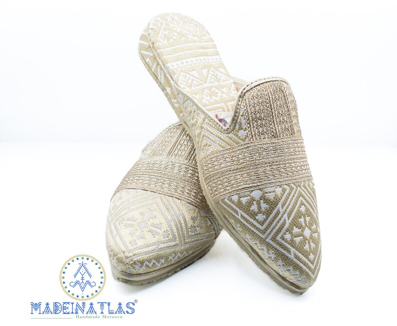 May include: A pair of gold and white patterned slippers with a pointed toe. The slippers have a woven strap across the top and a geometric pattern.