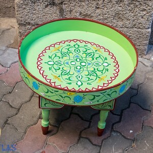 May include: A green painted wooden table with a round top and three legs. The top is decorated with a colorful floral design.
