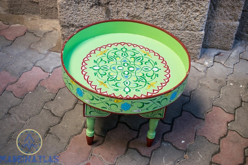 May include: A green painted wooden table with a round top and three legs. The top is decorated with a colorful floral design.