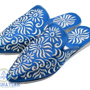 May include: A pair of blue embroidered slippers with a white floral pattern. The slippers are made in Morocco and have a pointed toe.