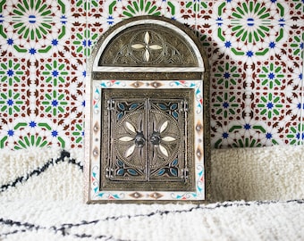 May include: A decorative mirror with a carved wooden frame and intricate metalwork. The frame is painted in shades of white, red, and blue. The mirror is set against a background of colorful Moroccan tiles.