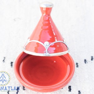 May include: A red ceramic tagine with a silver lid and decorative trim. The tagine is a traditional Moroccan cooking pot.