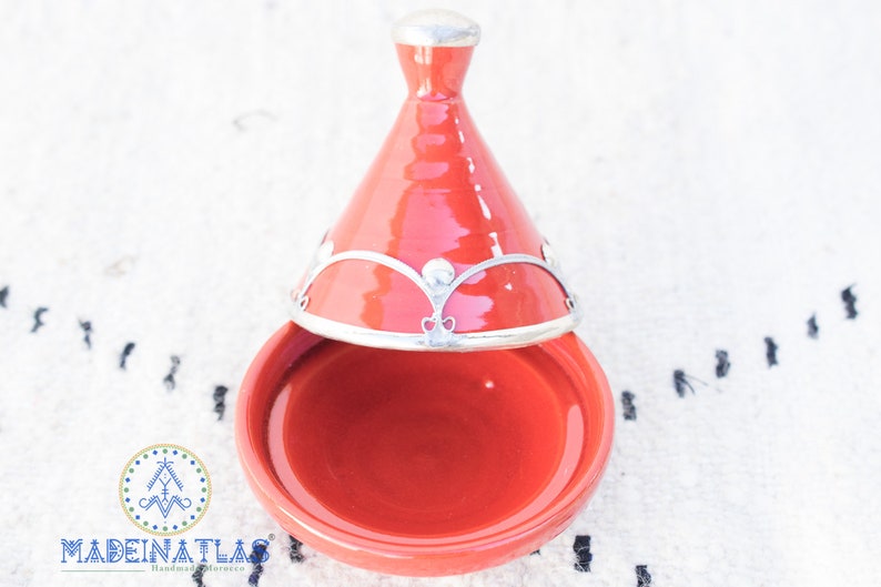 May include: A red ceramic tagine with a silver lid and decorative trim. The tagine is a traditional Moroccan cooking pot.