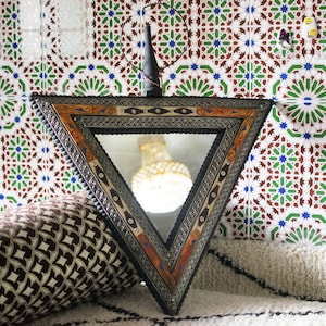 May include: A triangular mirror with a decorative, ornate frame in shades of brown, gold, and silver. The mirror is set against a background of white tiles with intricate green and brown geometric patterns.