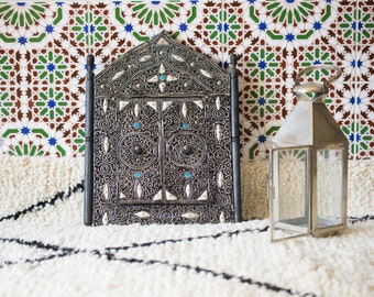 May include: A silver Moroccan-style mirror with intricate carvings and turquoise accents sits on a white fluffy rug in front of a wall with colorful geometric tiles.