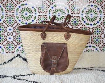 May include: A large, woven straw tote bag with brown leather handles and a brown leather pocket on the front. The bag is sitting on a white rug with black stripes.
