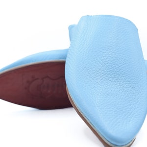 Puede incluir: Un par de zapatillas de cuero azul claro con punteras puntiagudas. Las zapatillas tienen una superficie texturizada y un ribete blanco alrededor de los bordes.