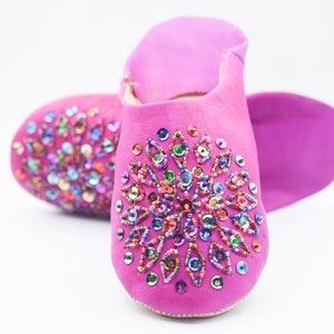 Può includere: Un paio di pantofole in pelle rosa con decorazioni di paillettes colorate. Le pantofole hanno una punta a punta e una suola piatta.