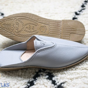 May include: A pair of gray leather slippers with a pointed toe and a brown leather sole. The sole has the text "Sandaliette" and "X For M" printed on it.