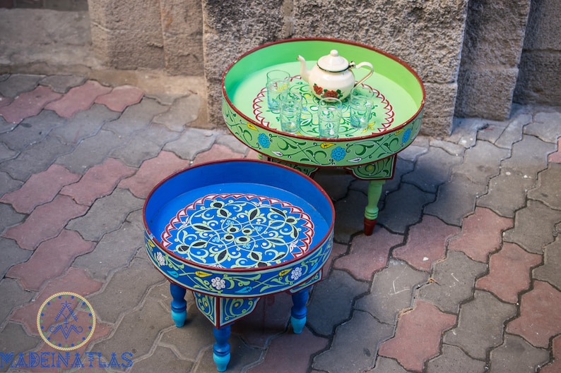May include: Two round, painted wooden tables with intricate floral designs. The larger table is green with a white teapot and glasses, while the smaller table is blue with a red and yellow border.