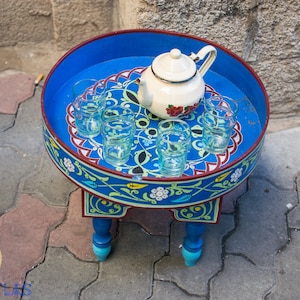 May include: A blue and red painted wooden table with intricate floral designs. The table has a round top with a white teapot and six clear glasses.