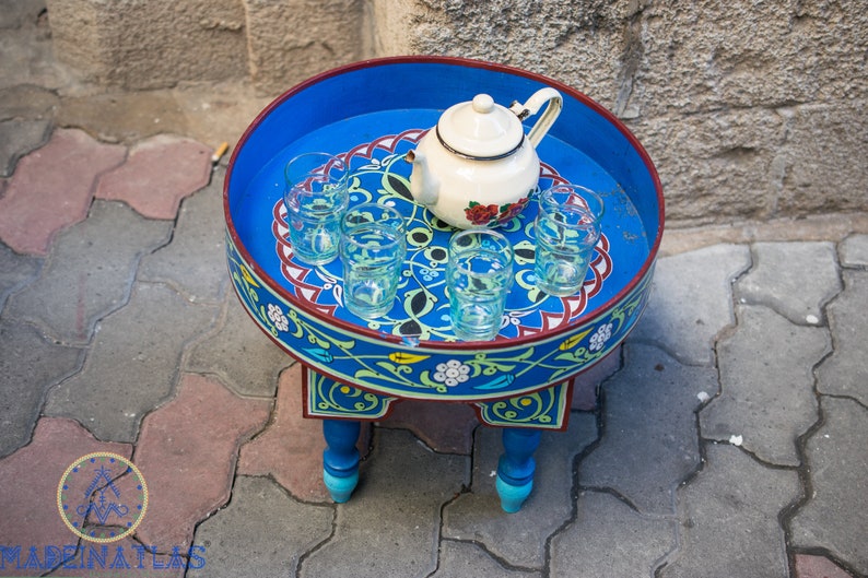 May include: A blue and red painted wooden table with intricate floral designs. The table has a round top with a white teapot and six clear glasses.