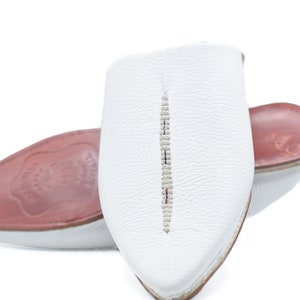May include: White leather slippers with a pointed toe and a decorative stitch down the center. The slippers have a red leather insole.