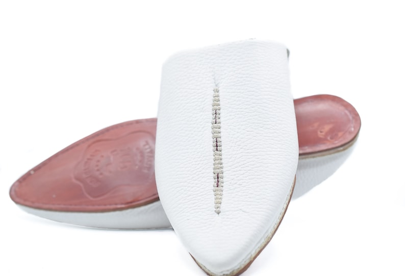May include: White leather slippers with a pointed toe and a decorative stitch down the center. The slippers have a red leather insole.