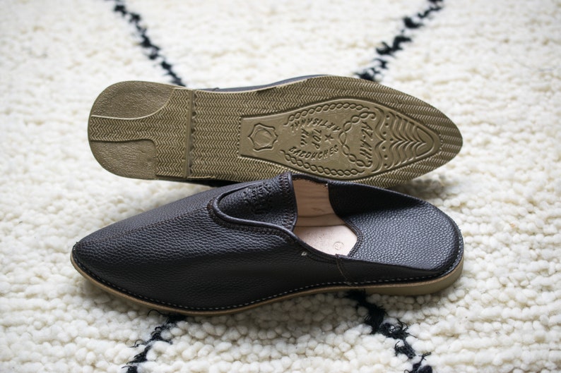 May include: A pair of black leather slippers with a brown sole. The sole has the text "TAYYELL" and "TOP" in a circle.