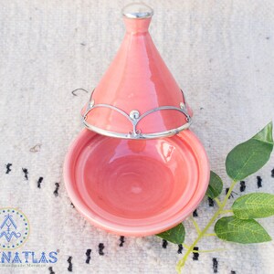 May include: A pink ceramic tagine with a silver lid and a silver trim around the top. The tagine is sitting on a white rug with black designs.