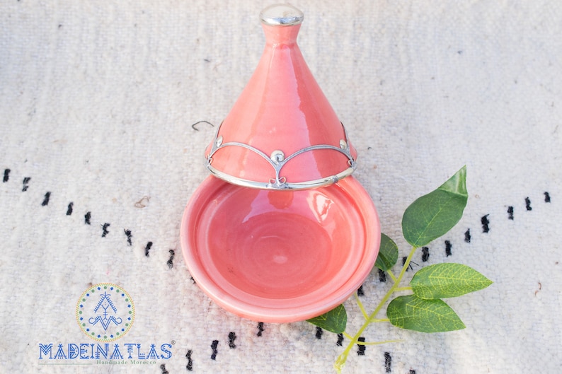 May include: A pink ceramic tagine with a silver lid and a silver trim around the top. The tagine is sitting on a white rug with black designs.
