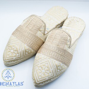 May include: Pair of gold embroidered slippers with a pointed toe and a woven strap across the top. The slippers have a white background with a geometric pattern.