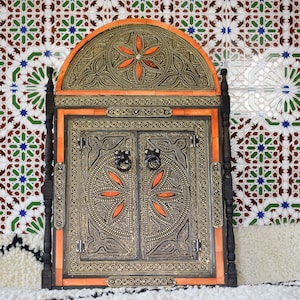 May include: A decorative wooden framed mirror with an arched top and intricate metalwork. The mirror has two doors with ornate metal designs and a central floral motif. The mirror is set against a background of colorful patterned tiles.