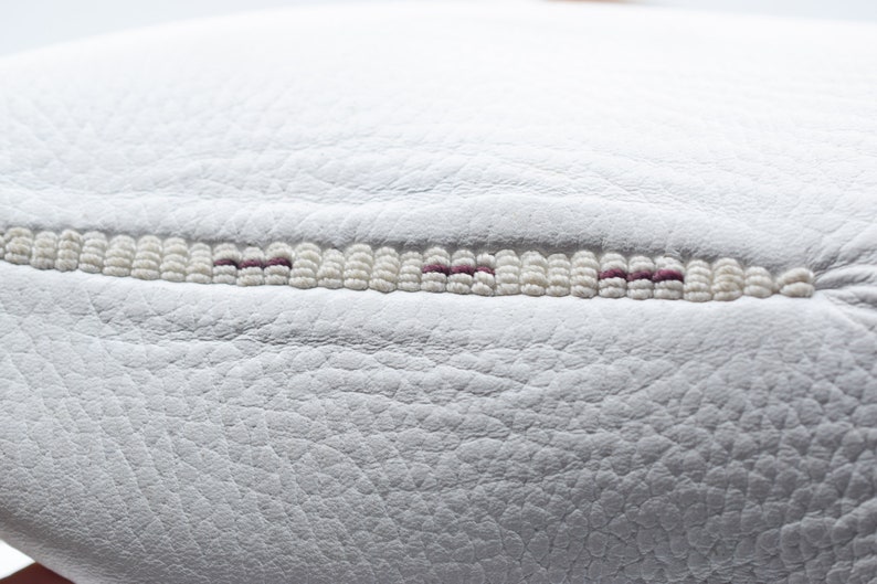 May include: Close-up of a white leather surface with a decorative stitch detail. The stitch is made of white beads with a thin line of purple thread running through the center.